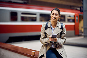 Junge Frau wartet am Bahngleis mit Smartphone in der Hand